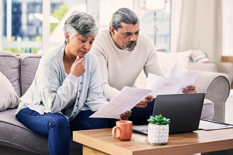 An older couple at home reviewing plan paperwork together with a laptop open on the table.