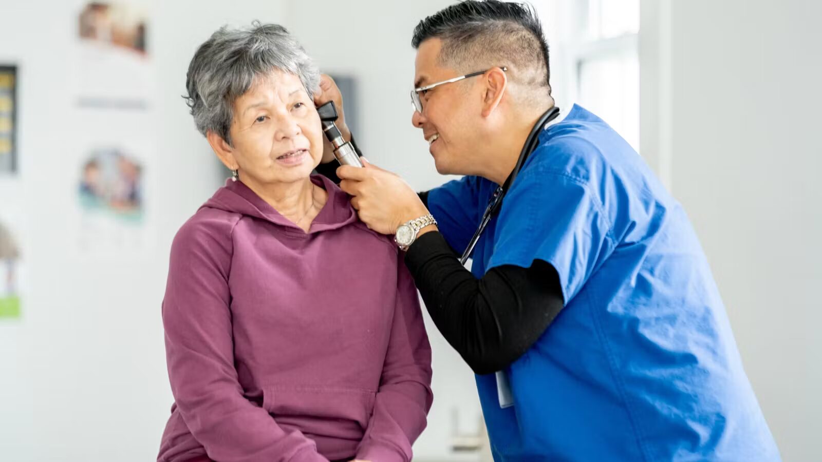 A doctor in blue scrubs looks into an older woman's ear with an otoscope.