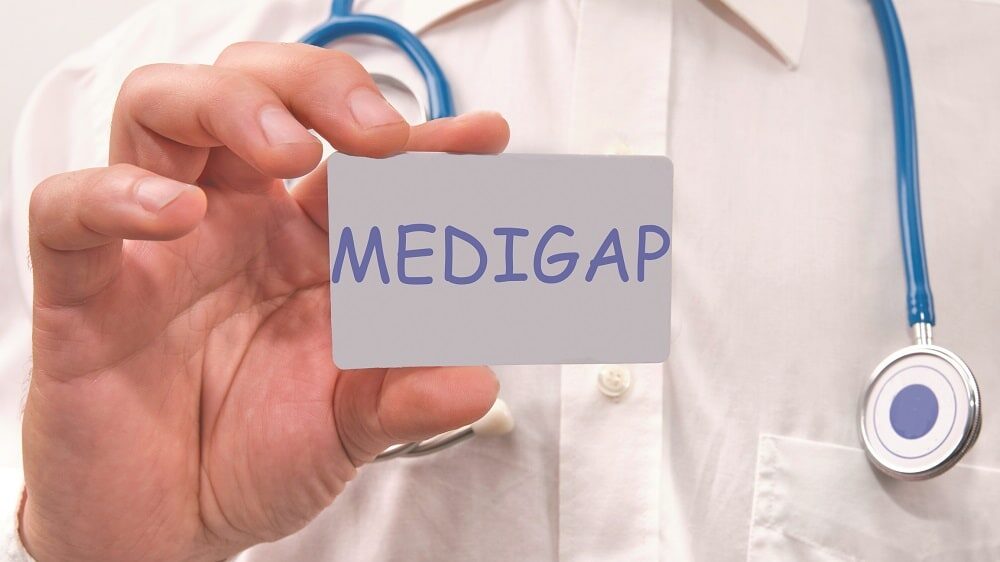 A doctor in a white coat with a blue stethoscope holds up a card reading Medigap.