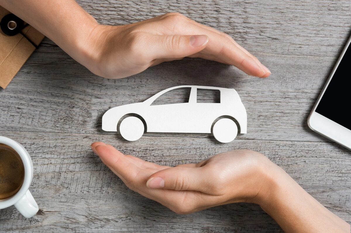 Two hands framing a white paper cutout of a car on a grey wooden table, beside a coffee cup and a phone.