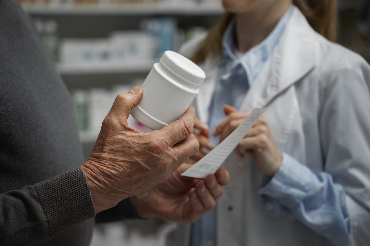 Close up of an older man's hand holding a white pill bottle while a pharmacist points at a printed list.