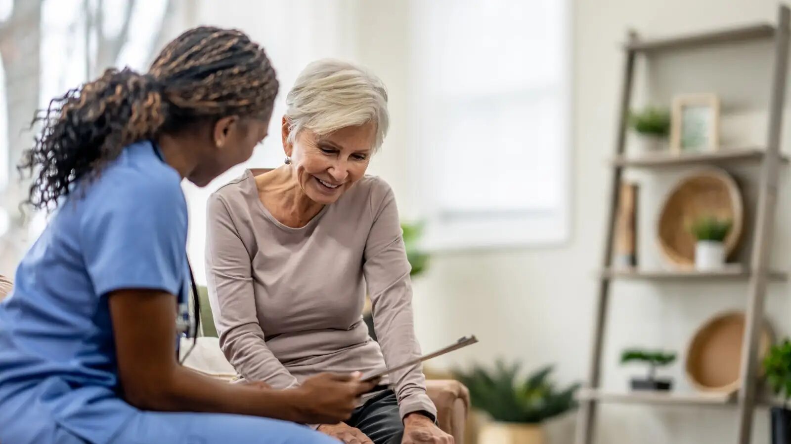 A nurse in blue scrubs and an older woman look over a clipboard together at home.