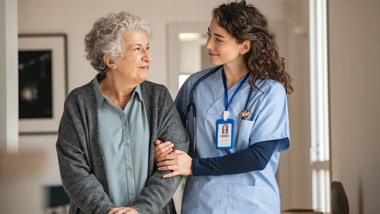 A nurse in blue scrubs walks arm in arm with a smiling older woman in a grey cardigan.