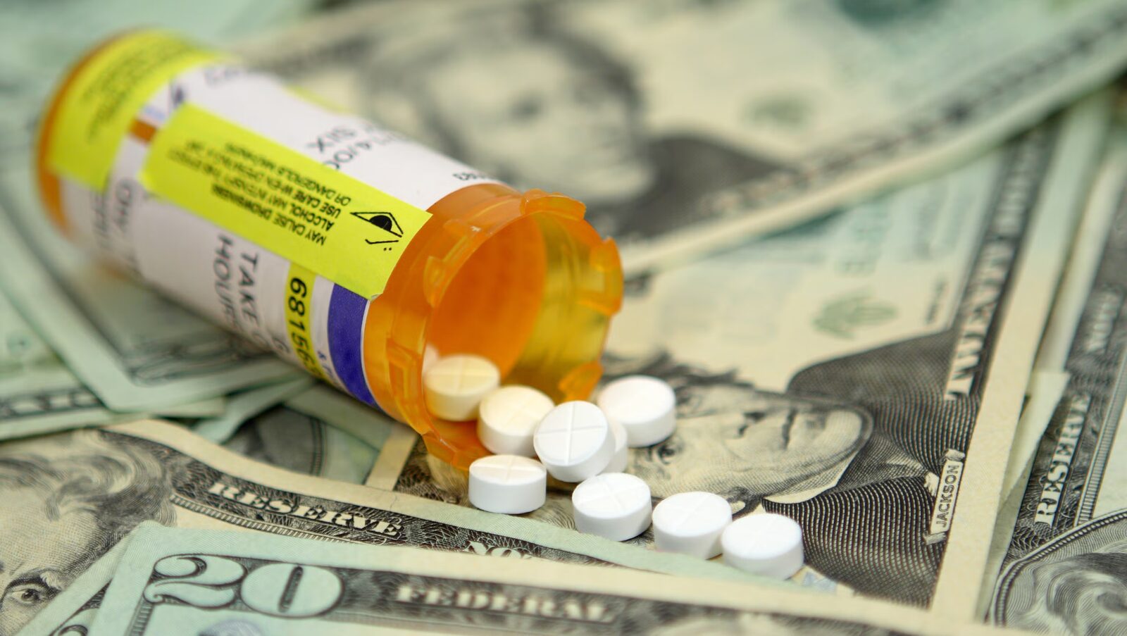 An orange prescription bottle lies open with white pills spilling out across twenty dollar bills.