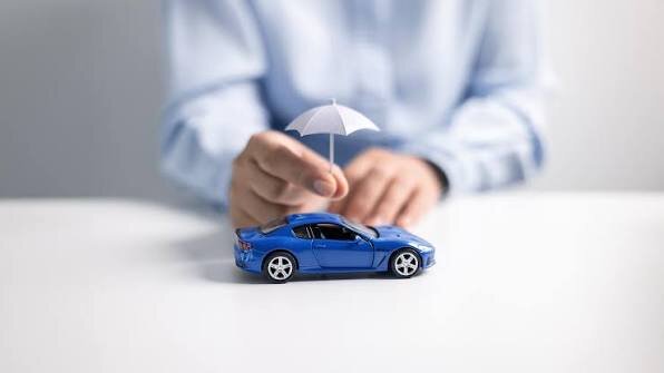 A person holding a tiny white umbrella over a blue toy car on a white desk.