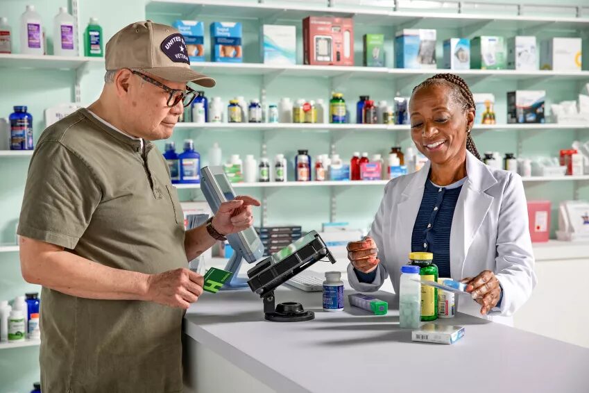 A man in a veteran's cap reads a medication box at a pharmacy counter while the pharmacist rings him up.